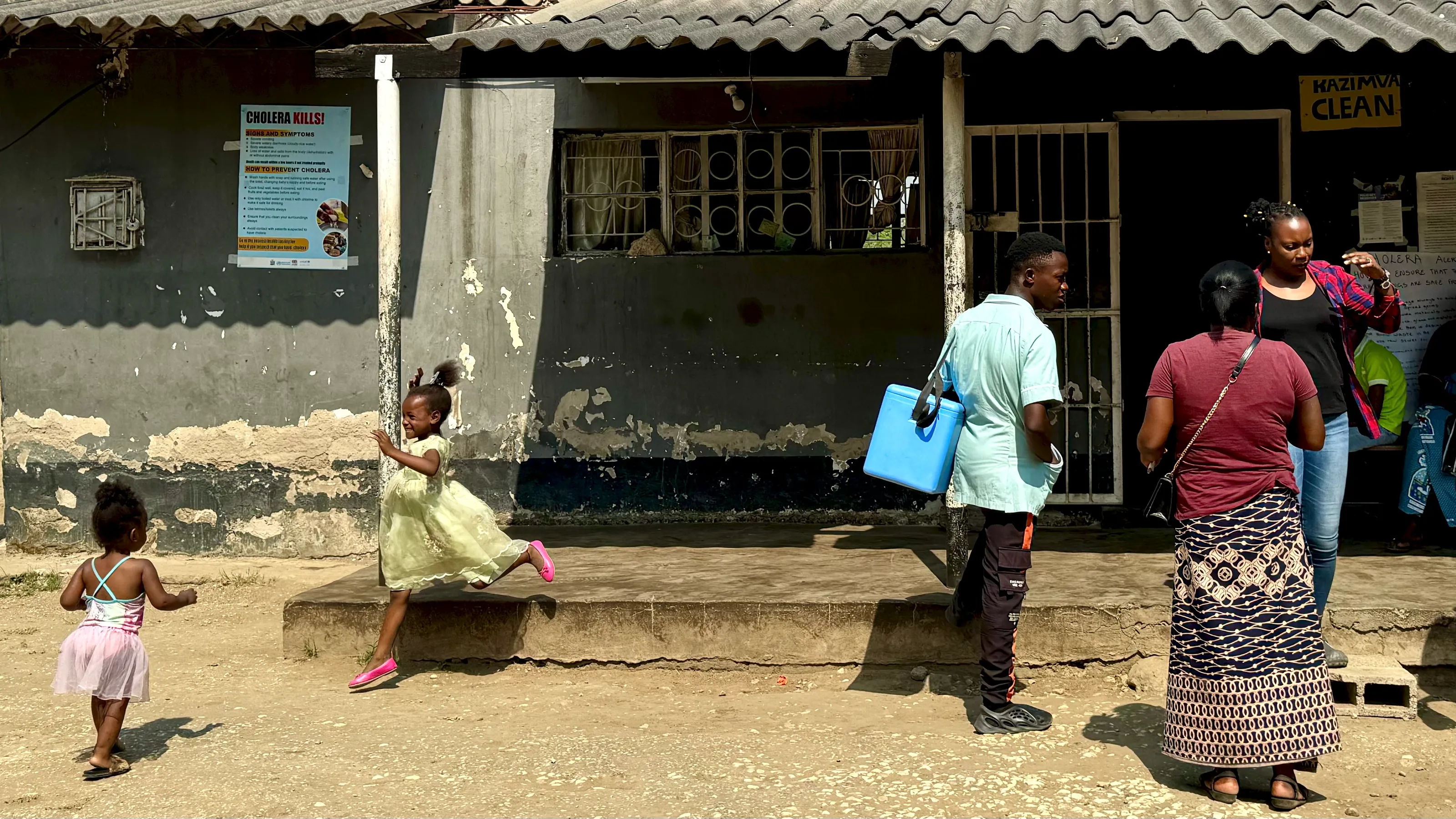 A frame from Polio’s Last Mile: outside a rural health post, a health worker carries a blue vaccine cold box while children play in the yard
